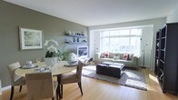 This well-appointed single-floor space showcases a thoughtfully designed open-plan layout that seamlessly connects the living and dining areas. The living room is anchored by a large bank of floor-to-ceiling windows that flood the space with natural light and offer views of the surrounding cityscape. A contemporary sofa with bold geometric-patterned accent pillows sits atop a textured area rug, complemented by a sleek dark coffee table that adds contrast to the light-toned interior. Decorative floating shelves on the feature wall display an artful arrangement of vases and sculptures, adding character and depth to the space.

The dining area features a round light-wood table paired with upholstered chairs, creating an intimate setting ideal for entertaining. Fresh orchids and greenery bring a natural, organic element to the room. Light hardwood flooring runs throughout, enhancing the sense of openness and warmth. The neutral color palette — soft greys, warm taupes, and crisp whites — creates a calm and cohesive ambiance throughout. Together, the architectural details and curated furnishings give the space a polished, urban-contemporary feel that is both inviting and functional.