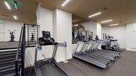 This fitness center is designed to support a comprehensive workout experience within a single-level layout. The space is divided into distinct functional zones, each outfitted with professional-grade equipment. The cardio area houses multiple treadmills with individual digital consoles, a row of elliptical trainers, StairMaster step mills, and stationary bikes, all arranged to allow comfortable spacing between users. A dedicated strength training zone features multiple cable machines, weight stations, and a Concept2 rowing machine, providing a full range of resistance training options.

The interior design is clean and modern, with crisp white walls contrasting against warm tan-toned ceilings. Rubber-speckled flooring runs throughout, offering durability and impact absorption. Recessed linear LED light fixtures illuminate the space evenly, creating a bright and focused atmosphere. Structural columns are incorporated into the layout without impeding usability, and large mirrors are strategically placed to expand the visual depth of the space. A cable railing system separates a lower-level strength area, adding a subtle architectural detail. The overall ambiance is polished and functional, reflecting a professionally designed facility suited for diverse fitness needs.