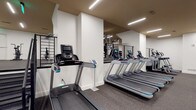 This fitness center is designed to support a comprehensive workout experience within a single-level layout. The space is divided into distinct functional zones, each outfitted with professional-grade equipment. The cardio area houses multiple treadmills with individual digital consoles, a row of elliptical trainers, StairMaster step mills, and stationary bikes, all arranged to allow comfortable spacing between users. A dedicated strength training zone features multiple cable machines, weight stations, and a Concept2 rowing machine, providing a full range of resistance training options.

The interior design is clean and modern, with crisp white walls contrasting against warm tan-toned ceilings. Rubber-speckled flooring runs throughout, offering durability and impact absorption. Recessed linear LED light fixtures illuminate the space evenly, creating a bright and focused atmosphere. Structural columns are incorporated into the layout without impeding usability, and large mirrors are strategically placed to expand the visual depth of the space. A cable railing system separates a lower-level strength area, adding a subtle architectural detail. The overall ambiance is polished and functional, reflecting a professionally designed facility suited for diverse fitness needs.
