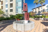Located at 500 Race St in San Jose, California, this generously sized outdoor courtyard serves as a vibrant communal gathering space nestled within a modern residential complex. The ground-level space is defined by its eye-catching geometric paver design, laid in a contrasting two-tone pattern that extends across the expansive open-air plaza.

A striking sculptural installation anchors one end of the courtyard, surrounded by a decorative stone planter that adds an artistic focal point to the space. Nearby, a central fountain feature provides a tranquil ambiance, framed by a curved architectural structure with glass entry doors. The courtyard is richly landscaped with a diverse mix of palms, ornamental trees, cycads, agaves, and native plantings arranged in raised stone-walled garden beds, creating a lush, resort-like atmosphere.

Outdoor dining areas are furnished with tables and chairs shaded by large umbrellas, ideal for casual gatherings and al fresco meals. A dedicated outdoor kitchen area with a stainless steel grill and a pergola overhead adds to the entertainment functionality of the space. Surrounded on multiple sides by the multi-story residential buildings, the courtyard benefits from an open sky above, abundant natural light, and a sheltered, private feel throughout.