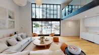 This single-floor space showcases a striking contemporary design defined by its dramatic double-height ceilings and an open, airy atmosphere. The main living area is anchored by expansive floor-to-ceiling, steel-framed windows and sliding glass doors that frame direct views of an outdoor pool and lounge area, flooding the interior with abundant natural light.

A glass-railed mezzanine overlooks the main floor, adding architectural depth and visual interest to the space. The warm, wide-plank hardwood flooring runs throughout, complementing the clean white walls, one of which features elegant vertical ribbed paneling that adds subtle texture to the interior.

The living area is generously proportioned, comfortably accommodating a large sectional sofa, woven accent chairs, and a round wooden coffee table arranged atop a natural fiber area rug. Pendant globe light fixtures provide soft ambient lighting.

Along one wall, a sleek built-in kitchenette features handle-free white cabinetry, a black matte faucet, and a minimalist countertop, maintaining the space's cohesive modern aesthetic. Recessed lighting and an upper mezzanine level with its own black-framed railing complete the sophisticated, loft-inspired design.