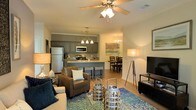 This single-floor residence offers a thoughtfully designed open-concept layout that seamlessly connects the living, dining, and kitchen areas into one inviting, cohesive space. The living room is anchored by warm hardwood-style flooring, a ceiling fan with integrated lighting, and crown molding that adds a refined architectural touch. Comfortable seating, layered rugs, and stylish décor accents create a welcoming and relaxed ambiance.

The kitchen is a standout feature, showcasing gray cabinetry, stainless steel appliances — including a refrigerator, microwave, and range — and a peninsula with bar-height seating, ideal for casual dining or entertaining. A pendant chandelier overhead adds warmth and character to the kitchen zone.

Just beyond, a dedicated dining area provides space for a full dining table and chairs, illuminated by an elegant chandelier that echoes the kitchen's lighting style. A piece of wall art adds a decorative focal point to this corner of the home.

Throughout the space, neutral wall tones and consistent flooring create a harmonious flow, while thoughtful lighting — including floor lamps and overhead fixtures — ensures a warm, well-lit atmosphere at every angle. The overall design balances functionality with comfort, making it a practical yet stylish living environment.