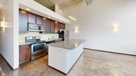 Located at 675 N Main St in downtown Winston-Salem, NC, this two-bedroom, two-bathroom unit offers an industrial loft aesthetic with thoughtful modern finishes throughout. The open living area makes a strong first impression with soaring ceilings, exposed ductwork, and polished concrete floors that lend an authentic urban character to the space. Architectural columns add structural interest to the open floor plan, while wall sconces provide warm ambient lighting.

The kitchen is well-appointed with rich dark wood cabinetry, stainless steel appliances including a range and refrigerator, a stainless microwave, and granite countertops that extend to a peninsula island — ideal for casual dining or meal prep. The open design connects the kitchen seamlessly to the living area, enhancing the spacious feel.

Both bedrooms continue the polished concrete flooring and crisp white walls, offering clean, versatile spaces. The primary bedroom features direct access to a dedicated closet and an en-suite bathroom. The bathrooms are finished with tile flooring and granite-topped vanities; one includes a full tub/shower combo, while the second offers a pedestal sink and clean, minimalist design. A hallway connects the private areas of the unit efficiently.