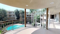 This single-floor space showcases a well-designed covered outdoor walkway and a striking curved balcony that together form a seamless transitional area within a larger multi-building complex. The corridor is defined by textured stucco ceilings and walls finished in warm, neutral tones, with a series of structural columns lining the open side and light blue metal railings providing safety along the length of the walkway. A skylight integrated into the ceiling allows natural light to filter in, brightening the passage throughout the day. The flooring throughout is a smooth, neutral-toned surface that complements the overall palette of the space. At the end of the corridor, the walkway opens onto a generously sized curved balcony that serves as the architectural highlight of the space. The balcony offers sweeping, unobstructed views of a resort-style courtyard below, featuring a sparkling swimming pool surrounded by tall palm trees and dense, mature landscaping. The surrounding multi-story buildings frame the view, creating a sense of enclosure while the open sky and greenery maintain an airy, sun-drenched ambiance. Together, the corridor and balcony provide a functional yet visually engaging outdoor experience.