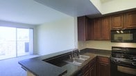 This single-floor residence offers a practical and comfortable layout designed for everyday living. The kitchen is a standout feature, showcasing rich dark wood cabinetry that provides generous storage space, paired with deep dark granite countertops that lend a grounded, classic aesthetic. A double-basin stainless steel sink sits at the center of the kitchen workspace, offering both utility and convenience. The kitchen opens into an adjacent living area, creating a connected and sociable flow between spaces. The living area is finished with wall-to-wall carpeting, contributing a warm and inviting atmosphere. Natural light filters into the space through a covered window, brightening the interior and enhancing the overall sense of openness. The ceilings feature a textured finish throughout, adding a layer of visual depth to the space. The neutral wall tones throughout provide a versatile backdrop, allowing for a wide range of interior styling choices. Overall, the residence presents a clean, functional environment with classic finishes that balance practicality with comfort.