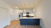 Located at 2 N Main St in Mount Prospect, Illinois, this single-floor unit offers a thoughtfully designed open-concept layout with contemporary finishes throughout. The kitchen serves as the centerpiece of the living space, showcasing a striking navy blue island with a white quartz countertop, an undermount sink, and sleek white upper cabinetry complemented by dark lower cabinets. Stainless steel appliances — including a French door refrigerator, gas range, and built-in microwave — complete the modern kitchen ensemble. Pendant lighting with gold-toned fixtures adds a warm, refined touch above the island. Light-toned hardwood-style flooring runs continuously throughout the unit, creating a cohesive and airy feel. The entry area provides convenient closet storage and direct access to the main living spaces. The living area benefits from natural light streaming through a large sliding glass door, offering outdoor views of surrounding greenery. A separate bedroom features a corner window arrangement with dual windows framing tree-lined views, filling the room with abundant natural light. The clean, minimalist design aesthetic, bright white walls, and open layout give the space a fresh and welcoming ambiance well-suited for modern living.