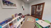 Kids Cave is a thoughtfully designed children's play area that provides a fun and functional environment for young ones. The space features soft carpet flooring throughout, creating a comfortable surface for play. A colorful activity rug anchors the center of the room, adding a playful focal point to the layout.

Along one wall, a low white shelving unit offers generous storage for toys, books, and play accessories, keeping the space organized and accessible for children. A child-sized table with bright teal and green chairs provides a dedicated spot for arts, crafts, or activities. A wall-mounted flat-screen TV adds an entertainment element, making this a truly versatile space.

The room is finished with light gray walls, warm wood baseboards, and recessed ceiling lighting that keeps the area bright and welcoming. A cozy bean bag chair tucked in the corner adds a relaxed seating option. Rich, dark wood-trimmed French doors with glass panes serve as the primary entry point, offering a glimpse into an adjacent fitness area beyond. An additional solid wood panel door provides further access within the space. The combination of practical storage, engaging furnishings, and quality finishes makes Kids Cave an ideal retreat designed with children in mind.