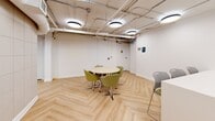 Located at 70 Spadina Rd in Toronto's Annex neighbourhood, this well-appointed amenity space is thoughtfully divided into two distinct zones, each designed to support different modes of gathering and relaxation. The entire area is unified by striking light-toned herringbone-patterned flooring that adds warmth and visual interest throughout.

The first zone serves as a combined kitchenette and casual dining area. A sleek white countertop with a built-in sink runs along one wall, accompanied by modern bar-height seating. A round table with contemporary green upholstered chairs provides additional seating, while a tall potted plant and a framed coastal artwork contribute a fresh, welcoming atmosphere. Exposed ceiling infrastructure, including conduit and ductwork, lends an industrial edge that contrasts with the clean white walls.

The second zone is a comfortable lounge area anchored by a bold blue geometric-tiled accent wall that immediately draws the eye. A blue leather sofa, a vibrant orange swivel chair, and a low wooden coffee table create an inviting conversational setting. A wall-mounted flat-screen TV adds a functional element. Circular pendant lights illuminate both areas with a warm, even glow, tying the space together with a cohesive and contemporary aesthetic.