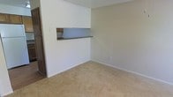 This two-story space offers a straightforward and functional layout designed for everyday living. The main living area is finished with wall-to-wall beige carpeting, creating a warm and neutral foundation throughout the room. Smooth, light-colored walls complement the textured ceiling, contributing to a clean and understated aesthetic.

A notable architectural feature is the built-in pass-through window connecting the living area to the kitchen. This opening, accented by a dark countertop ledge and wooden cabinetry visible beyond, allows for easy communication and service between the two spaces. The design promotes an open feel while maintaining a defined separation between the living and kitchen areas.

The living room provides ample open floor space, making it adaptable for a variety of furniture arrangements. Subtle details such as clean baseboards and a standard electrical outlet along the wall reflect a practical, well-maintained interior. The overall ambiance is calm and neutral, offering a blank canvas that accommodates a range of interior styles. The two-floor configuration suggests additional living or sleeping space on the upper level, adding to the overall functionality of the home.