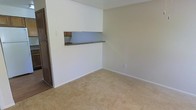 This two-story space offers a straightforward and functional layout designed for everyday living. The main living area is finished with wall-to-wall beige carpeting, creating a warm and neutral foundation throughout the room. Smooth, light-colored walls complement the textured ceiling, contributing to a clean and understated aesthetic.

A notable architectural feature is the built-in pass-through window connecting the living area to the kitchen. This opening, accented by a dark countertop ledge and wooden cabinetry visible beyond, allows for easy communication and service between the two spaces. The design promotes an open feel while maintaining a defined separation between the living and kitchen areas.

The living room provides ample open floor space, making it adaptable for a variety of furniture arrangements. Subtle details such as clean baseboards and a standard electrical outlet along the wall reflect a practical, well-maintained interior. The overall ambiance is calm and neutral, offering a blank canvas that accommodates a range of interior styles. The two-floor configuration suggests additional living or sleeping space on the upper level, adding to the overall functionality of the home.