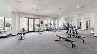 Located at 5942 Factory Shops Boulevard in Ellenton, Florida, this fitness center offers a comprehensive, single-floor layout designed to accommodate a wide range of workout needs. The main exercise room is expansive and generously lit by large windows that frame views of the surrounding landscape, including glimpses of a pool and palm trees, creating an energizing and open atmosphere. Soaring double-height ceilings in key areas of the main floor amplify the sense of space, while crisp white walls and recessed lighting maintain a clean, modern aesthetic throughout. The exercise room features durable, textured carpet flooring and is outfitted with a wide array of equipment, including treadmills, ellipticals, stationary and spin bikes, stair climbers, cable machines, strength training machines, free weights, and weight benches. Floor-to-ceiling mirrors along multiple walls enhance the sense of depth and support proper form during workouts. A dedicated studio space branches off from the main area, finished with light wood-tone flooring and enclosed by sleek black-framed glass walls and doors. This room is equipped with resistance bands, stability balls, yoga mats, and a large display screen, making it ideal for group classes or individual training sessions. The hallway connects these areas and includes restroom access and a wall-mounted water fountain, ensuring full convenience for facility users.