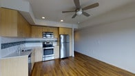 This single-floor space showcases an open-concept layout that seamlessly connects the kitchen and living area. The kitchen is well-appointed with stainless steel appliances, including a refrigerator, range with oven, and built-in microwave. Warm-toned wood cabinetry provides ample storage above and below the counter, complemented by a grey tile backsplash that adds a modern touch. Light-colored lower cabinets and countertops contrast nicely with the upper cabinetry, creating a balanced, contemporary aesthetic.

Rich, dark hardwood flooring extends throughout both the kitchen and the adjoining living area, adding warmth and continuity to the space. The living area is open and versatile, offering a blank canvas for various furniture arrangements. Recessed lighting in the kitchen ensures functional illumination, while a ceiling fan with a built-in light fixture in the living area provides both comfort and ambient light. A tray ceiling detail adds architectural interest overhead. The neutral wall tones throughout keep the space feeling bright, clean, and move-in ready.