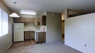 This single-floor space features an open layout connecting the kitchen and living area, offering a functional and straightforward design. The kitchen is equipped with medium-toned wood laminate cabinetry on both upper and lower sections, providing ample storage. White appliances, including a refrigerator, stove, and dishwasher, complement the light countertops and double-basin sink positioned beneath a fluorescent overhead light fixture. The kitchen flooring is hardwood-style, transitioning into carpet in the adjacent living area, which offers a comfortable, casual atmosphere. A partial wall divides the kitchen from the rest of the space, creating a subtle sense of separation while maintaining the open feel. The textured popcorn ceiling is consistent throughout. A bathroom is visible through a doorway at the rear, indicating a practical, compact layout. The neutral beige and tan color palette throughout the space provides a clean, understated aesthetic well-suited for everyday living.
