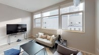 This contemporary single-floor residence offers a well-designed layout that balances modern aesthetics with functional living. The open-concept kitchen is a standout feature, showcasing white shaker cabinetry, light quartz countertops, a herringbone tile backsplash, and a full suite of stainless steel appliances. A central island with seating provides both additional prep space and a casual dining area, while industrial-style pendant lighting adds character overhead. Light wood-look flooring flows throughout the main living areas, contributing to a clean, cohesive aesthetic. The living room is bathed in natural light through a wide bank of windows that frame elevated urban cityscape views, lending an airy, connected feel to the space. Exposed ductwork on the ceiling adds an industrial design element that complements the modern finishes. The two bedrooms offer a calm retreat, with carpeted floors, subdued neutral tones, and high ceilings with exposed mechanical details. The bathroom is thoughtfully appointed with white cabinetry, a well-lit vanity mirror, and a tall storage cabinet for added convenience. A stacked washer and dryer unit is housed in a dedicated nook, providing in-unit laundry. The entryway features a contemporary door and a smart thermostat, reflecting the home's modern, tech-forward sensibility throughout.
