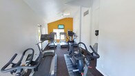 This fitness center is housed in a single-story space distinguished by a vaulted ceiling that lends an open, airy feel to the room. The ceiling is equipped with a ceiling fan to help maintain airflow during workouts. A bold amber accent wall at the far end of the room serves as a focal point, complementing the otherwise neutral white walls and creating an energizing atmosphere.

The space is outfitted with a variety of cardio equipment, including multiple treadmills, elliptical machines, and a stationary bike, all arranged to allow comfortable use. Dark, durable flooring runs throughout, providing a practical surface suited to exercise. A wall-mounted television is positioned above the accent wall, offering entertainment options for users during their workouts.

Natural light enters through windows along the side wall and a glass door at the far end, offering views of the surrounding outdoor area. A sanitation station is mounted on the wall, supporting hygiene between equipment use. The overall layout is efficient and functional, making good use of the available space while providing essential cardio fitness amenities.