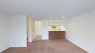 This single-floor space presents an open and functional layout designed for comfortable everyday living. The main living area is finished with wall-to-wall neutral-toned carpeting, lending a soft, understated feel to the space. White walls throughout keep the interior bright and clean, while recessed lighting in the ceiling provides focused illumination across the room.

A defining architectural feature is the half-wall peninsula that separates the living area from the kitchen, creating a pass-through that maintains an open connection between the two spaces. The kitchen, visible beyond the partition, features white cabinetry and appliances, contributing to the cohesive, light color palette. A range hood is mounted above the cooking area, and the kitchen counter offers practical workspace.

Toward the rear of the space, a hallway leads to additional areas and is flanked by white bi-fold closet doors, suggesting generous built-in storage capacity. A window with horizontal blinds at the end of the hallway introduces natural light into the corridor. The overall ambiance is clean, neutral, and move-in ready, offering a versatile canvas for personalization.