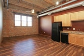 This single-floor unit showcases a compelling industrial-loft aesthetic, blending raw architectural elements with practical living spaces. The open-concept main living and kitchen area is defined by soaring wood-beam ceilings with exposed metal ductwork running overhead, lending a warehouse-inspired character throughout. Dark hardwood-style flooring extends across the main living space, complementing bold, deep-toned accent walls and structural wood columns that punctuate the room. A ceiling fan provides airflow in the generous living area, which also benefits from natural light through a window framed by original brick accent walls.

The kitchen is well-appointed with light maple cabinetry reaching toward the ceiling, a double-basin sink, and a full suite of black appliances including a refrigerator, dishwasher, microwave, and range. Laminate countertops provide ample prep space.

The bedroom areas feature carpeted flooring, neutral taupe walls, and exposed overhead ductwork consistent with the unit's industrial character. Sliding barn-style doors add a functional, space-conscious design detail. The bathroom includes a tub-shower combination, a wood-toned vanity with a single sink, a large mirror, and wood-look vinyl flooring, maintaining a clean and cohesive finish throughout.