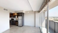 This contemporary single-floor unit showcases a clean, modern aesthetic throughout, with polished concrete flooring and neutral-toned walls creating a cohesive and understated palette. The kitchen is a standout feature, outfitted with dark espresso cabinetry that extends to the ceiling for ample storage, a full suite of stainless steel appliances including a refrigerator, electric range, microwave, and dishwasher, and a single-basin sink. Track lighting illuminates the kitchen workspace, while the open layout connects seamlessly to the living area, which benefits from large sliding glass doors that invite natural light and provide access to an outdoor space. The bedroom is a bright, well-proportioned room with large windows allowing generous sunlight and views to the outside. Two bathrooms mirror each other in design, each featuring a frameless sliding glass-enclosed walk-in shower with large-format white tile surround, a wall-mounted vanity with granite countertop and undermount sink, a full-width mirror, and dark cabinetry with open shelving for storage. The consistent use of concrete flooring, dark wood-tone finishes, and minimalist fixtures throughout the unit creates a unified, contemporary living environment that is both functional and visually refined.