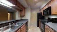 This single-story space offers a straightforward, functional layout designed for comfortable everyday living. The main living area features neutral-toned walls and wall-to-wall carpet in a warm beige hue, creating a cohesive and understated aesthetic throughout. A structural column and angled wall partition define the space, adding architectural interest while guiding the flow between rooms. Ceiling-mounted HVAC vents are integrated throughout, and standard white baseboards provide a clean finish along the floors.

A hallway corridor connects the living area to additional rooms, with a glimpse of natural light visible at the far end, suggesting windows in the adjoining spaces. The kitchen is a galley-style layout, featuring rich medium-toned wood cabinetry that extends to the upper walls, providing ample storage. Dark laminate countertops run the length of the kitchen, paired with a stainless double-basin sink. Black appliances, including a refrigerator, electric range, built-in microwave, and dishwasher, are neatly integrated. Light tile flooring in the kitchen contrasts with the carpeted living areas, clearly delineating the two zones. Track lighting overhead adds focused illumination throughout the kitchen.