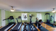 The Parc Bordeaux fitness center is a thoughtfully designed, single-floor facility that combines functionality with an energizing atmosphere. The space is immediately welcoming, featuring warm hardwood floors that stretch throughout, vibrant green walls, and crown molding that adds a refined architectural touch. Natural light pours in through multiple windows adorned with blinds, as well as arched glass-panel entry doors that enhance the open, airy feel of the room.

The cardio section is well-stocked with treadmills, elliptical trainers, and a recumbent bike, all positioned along rubber floor mats to ensure safety and stability. The strength training area includes a multi-functional cable machine, a weight bench with free weights, exercise balls, and step platforms, offering a comprehensive range of workout options. A full-length mirror along one wall provides visual feedback during workouts and adds to the sense of spaciousness.

Motivational quotes are displayed prominently along the upper portion of the walls, reading phrases such as "Ability is what you're capable of doing. Motivation determines what you do. Attitude determines how well you do it," reinforcing an encouraging environment. Ceiling fans with overhead lighting ensure comfortable air circulation throughout. The layout is open and generously proportioned, providing ample room to move freely between equipment stations.