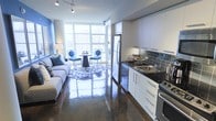 This single-floor space showcases a thoughtfully designed open-concept layout that seamlessly integrates the kitchen, living, and dining areas into one cohesive environment. The kitchen is outfitted with crisp white cabinetry, dark granite countertops, stainless steel appliances including a full-sized refrigerator, and a deep single-basin sink. A slate-blue tile backsplash adds a striking contrast to the white cabinetry, lending the kitchen a polished, contemporary aesthetic.

The highly reflective, dark-toned polished concrete floors extend throughout the entire space, contributing to a sleek, modern ambiance while amplifying the abundant natural light streaming in from the expansive floor-to-ceiling windows. These oversized windows frame urban views and serve as a defining architectural feature of the space.

The living area is furnished with a light-toned sectional sofa accented with blue throw pillows, while the compact dining area features a round table with elegant blue accent chairs. A patterned area rug anchors the dining and living zones, adding texture and warmth. Track lighting overhead provides additional illumination, complementing the natural light throughout.