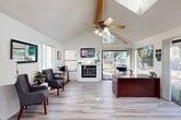 Located at 1420 Lake Blvd in Davis, California, this single-story office space offers a thoughtfully designed layout spread across four distinct areas. The welcoming reception area immediately impresses with its dramatic vaulted ceiling accented by exposed wooden beams, a ceiling fan, and a skylight that floods the space with natural light. Light wood-look flooring runs throughout, complementing the crisp white walls. A decorative fireplace serves as a focal point, topped by a wall-mounted TV, while a sliding glass door opens directly to an outdoor patio and pool area, further enhancing the bright, airy atmosphere. A reception desk anchors the front area, alongside comfortable seating for visitors. Adjacent to the reception area is a dedicated private office featuring large windows overlooking the outdoor pool, a substantial executive desk, and direct patio access via a sliding door. Two conference rooms provide functional meeting spaces, each furnished with a central conference table and ergonomic chairs. One room includes a whiteboard, framed artwork, and a leather sofa, while the other features a bulletin board and ample natural light through vertical-blind-covered windows. Rounding out the space is a laundry room with stacked washer and dryer units, a generous folding counter, and cheerful décor, adding a practical amenity to this versatile office.