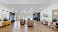 Located in Columbia, SC, this single-floor clubhouse features a generously sized, open-concept living area designed for comfort and community gathering. The space is anchored by a striking navy blue accent wall with a built-in fireplace and decorative mantel, creating a bold focal point that complements the otherwise bright, white interior.

Light hardwood-style flooring runs throughout, reflecting the abundant natural light that pours in through multiple sets of French doors. These glass doors line the exterior wall and offer direct views of the outdoor pool area, seamlessly blending indoor and outdoor living. Ceiling fans are mounted across the vaulted ceiling, adding both function and a casual, airy feel to the room.

The layout accommodates multiple seating arrangements, including plush sofas and accent chairs grouped around a central coffee table, as well as a dedicated workspace with a desk and seating near the entrance. A large sliding barn door in matte black adds an industrial-rustic touch, while a coffee and beverage station is tucked conveniently nearby.

Decorative elements such as hanging planters, potted greenery, framed artwork, and a sunburst mirror contribute to the space's warm, modern-rustic aesthetic. A separate glass-enclosed room, visible from the main area, appears to house fitness equipment, rounding out this well-appointed clubhouse.