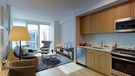 This well-appointed single-floor residence showcases a thoughtfully designed open-concept layout that seamlessly blends living and kitchen areas into one cohesive space. The interior is characterized by a clean, contemporary aesthetic, with crisp white walls and smooth ceilings that enhance the sense of openness and brightness throughout.

The living area is anchored by large floor-to-ceiling windows that flood the space with natural light and offer views of the surrounding urban environment. The room comfortably accommodates a seating arrangement centered around a sleek coffee table atop a patterned area rug, creating an inviting and functional gathering space. Light-toned hardwood flooring runs throughout, adding warmth and continuity to the overall design.

Adjoining the living area is a compact yet efficient kitchen, defined by rich warm-toned wood cabinetry that extends to the upper walls, providing generous storage. A stainless steel sink with a modern faucet and integrated dishwasher offer practical functionality, while light stone countertops complement the natural wood tones. The kitchen's recessed design gives it a refined, built-in appearance that contributes to the space's streamlined feel.

Overall, the residence strikes a balance between modern minimalism and warm, livable comfort, making it an ideal urban retreat.