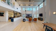 This single-story clubhouse in Mary Esther, Florida, is designed with a contemporary aesthetic that emphasizes open, airy living. The centerpiece of the space is a grand main hall with dramatic double-height ceilings and an expansive array of clerestory windows that flood the interior with natural light. Light-toned hardwood flooring runs throughout the main areas, complementing the crisp white walls and modern furnishings.

The lounge area features a comfortable seating arrangement with warm-toned accent chairs, a wall-mounted flat-screen television, and a stylish media console. A large overhead garage-style door opens the lounge toward the exterior, further enhancing the connection to the outdoors and views of the surrounding community. Adjacent to the lounge, a dedicated dining area accommodates a solid wood table with contemporary seating beneath floor-to-ceiling windows overlooking a pool and landscaped grounds.

The clubhouse also includes a fully equipped kitchen outfitted with bold teal cabinetry, granite countertops, mirrored tile backsplash, and stainless steel appliances. A glass-enclosed office or conference space with steel-framed partitions provides a functional workspace. Throughout, black-framed interior windows and modern wall sconces add refined architectural detail to this well-appointed community gathering space.