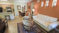 This single-story space showcases an open-concept layout that connects the living and dining areas, creating a cohesive and welcoming atmosphere. The interior is defined by a striking terracotta-orange accent wall that anchors the living area, adding warmth and visual depth to the space. Neutral cream-toned walls complement the bold color, maintaining a balanced and inviting palette throughout.

The living area features hardwood-style flooring that extends throughout, providing continuity between spaces. A white tufted sofa anchors the seating area, accompanied by a patterned accent chair and an oval glass-top coffee table resting on a multicolored area rug, adding texture and character to the room.

The dining area, visible from the living space, includes a dark wood dining table surrounded by rich brown upholstered chairs. A contemporary chandelier hangs above, providing focused ambient lighting. Decorative framed artwork adorns the walls, enhancing the curated feel of the space.

Adjacent to the dining area, a glimpse of the kitchen reveals light wood cabinetry. A hallway leads to additional rooms, suggesting a functional and well-organized floor plan throughout this single-level home.