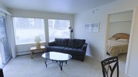 This single-floor space offers a comfortable and functional layout well-suited for everyday living. The living room serves as the central gathering area, featuring plush wall-to-wall carpeting that adds warmth underfoot. A large, tufted dark sofa anchors the seating area, complemented by an elegant oval glass-top coffee table with decorative metalwork legs. Two generously sized windows allow an abundance of natural light to fill the room, creating a bright and welcoming atmosphere. A side table with a ceramic table lamp adds a soft, ambient lighting option for evenings. Framed artwork on the wall provides a tasteful decorative accent. The walls are finished in a clean, neutral tone that enhances the sense of openness throughout the space. The room flows naturally toward an adjacent area, suggesting a practical and connected floor plan. Overall, the space presents a clean, understated aesthetic that balances simplicity with comfort, making it an inviting environment for both relaxing and entertaining.