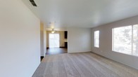 This single-floor residence offers a 2-bedroom, 2-bathroom layout designed with an open and functional floor plan. The living area features plush wall-to-wall carpeting and neutral-toned walls that create a clean, versatile backdrop. Natural light filters in through a side window, brightening the space comfortably. The layout flows seamlessly into an adjacent area with hard-surface flooring, visible through an open passage. That secondary space is equipped with a ceiling fan with light fixture and features a sliding glass door fitted with vertical blinds, providing access to an outdoor area and bringing in additional natural light. Dark cabinetry is visible in the background, suggesting a kitchen or dining area nearby. HVAC vents are positioned in both the ceiling and along the baseboards, ensuring climate control throughout. The open sightlines between the carpeted and hard-floor zones create a sense of connected, expansive living space. The overall ambiance is neutral and move-in ready, offering a straightforward and practical living environment suitable for a variety of lifestyles.