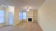 This single-floor residence offers a comfortable and well-appointed layout designed for everyday living. The living area serves as the heart of the home, drawing the eye immediately to a classic fireplace framed by a clean white mantel, which adds both warmth and a timeless focal point to the room. Flanking the fireplace are built-in shelves that provide generous display and storage space while contributing to the room's structured, polished aesthetic. Crown molding runs along the ceiling, lending a refined architectural detail that elevates the overall character of the space. Natural light fills the room through a set of bay windows, creating a bright and airy atmosphere that complements the neutral, warm-toned walls and plush carpeted flooring. A ceiling fan with integrated lighting is centrally positioned, ensuring comfort and illumination throughout the space. The soft, neutral color palette — from the cream-toned walls to the light beige carpet — creates a cohesive and inviting ambiance that is easy to personalize. The single-floor layout offers seamless accessibility and flow throughout the home, making it a practical and welcoming space suited for a variety of lifestyles.