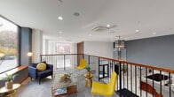 Located at 697 Bronx River Rd in Yonkers, NY, this thoughtfully designed two-floor building makes an immediate impression with its soaring double-height lobby. Large-format marble-look tile flooring stretches throughout the ground level, reflecting recessed lighting and creating an open, luminous atmosphere. Framed artwork adorns the charcoal-toned accent walls, while generous windows and glass entry doors invite abundant natural light and views of the surrounding greenery.

A wide staircase with a warm wood handrail and sleek black metal railings connects the ground floor to the upper level, where a spacious open lounge area awaits. The second floor features plush carpeting, bold yellow accent chairs, and a wood-toned bar-height workstation, all set against a backdrop of floor-to-ceiling windows that frame lush tree-lined street views. A built-in wood shelving unit adds warmth and function, while recessed and pendant lighting enhance the refined ambiance.

The first-floor corridors are bright and well-appointed, with polished marble-style tile, potted greenery, and subtle seating alcoves. A dedicated lounge area on the ground floor boasts soaring ceilings, expansive windows with tree views, and an elegant mix of contemporary furniture, making the entire space feel both welcoming and sophisticated.