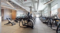 This large-scale fitness center in Denver, Colorado offers a comprehensive, well-appointed workout environment spread across a single, open floor plan. The facility's industrial-inspired architecture is defined by soaring ceilings with exposed ductwork and large-diameter pipes painted white, structural concrete columns, and linear pendant lighting that illuminates every zone of the facility.

The cardio area is outfitted with a generous row of treadmills, elliptical trainers, and stationary cycling equipment, all positioned to take advantage of the facility's oversized windows, which allow ample natural light and views of the surrounding Denver skyline. Rubber flooring throughout ensures durability and safety across all workout zones.

A dedicated strength training section houses an extensive array of selectorized machines and cable systems, organized for efficient traffic flow. A separate free weights area provides benches, barbell stations, dumbbells, and functional training equipment including plyo boxes and cable attachments. Warm terracotta and muted orange accent walls punctuate the space alongside exposed concrete, creating a modern yet energetic atmosphere. Full-length mirrors expand the sense of space visually while supporting proper training form. The facility's thoughtful layout, abundant equipment variety, and bright, welcoming design make it a fully equipped destination for all fitness levels.