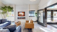 Located at 1410 Wendell Ave SE in Port Orchard, Washington, this thoughtfully designed single-floor amenity space offers a well-rounded collection of community-focused rooms connected by welcoming hallways. The entry corridor features warm wood plank flooring, soaring vaulted ceilings clad in natural tongue-and-groove wood paneling, and lush tropical greenery, setting an inviting tone throughout.

The lounge area is a standout gathering space, anchored by a floor-to-ceiling white brick accent wall with a built-in fireplace and wall-mounted television. Comfortable leather seating, soft area rugs, and abundant natural light from large windows create a relaxed, social atmosphere. Glass-paned doors lead directly to an outdoor area, enhancing the connection to the surrounding environment.

The community kitchen is fully appointed with light wood cabinetry, white quartz countertops, a classic subway tile backsplash, stainless steel appliances including a French door refrigerator and dishwasher, open shelving, and a dining bar with seating — ideal for communal use.

The fitness center is equipped with cardio machines, a cable multi-station, free weights, and mirrored walls, all beneath distinctive exposed wood beam ceilings and rubber flooring. A dedicated workstation room provides two adjustable desks and blue task chairs in a quiet, carpeted setting with exterior-facing windows.