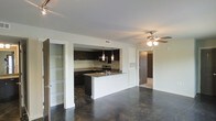 This single-floor space showcases a thoughtfully designed open-concept layout that seamlessly connects the living area to the kitchen. The polished concrete floors run throughout, lending an industrial yet refined aesthetic that anchors the entire space. The kitchen is a standout feature, outfitted with dark espresso cabinetry that contrasts sharply against light countertops and white walls. A peninsula with a built-in sink serves as both a functional workspace and a natural divider between the kitchen and living area. Pendant lights hang above the peninsula, providing focused task lighting and a decorative touch. Stainless steel appliances, including a microwave and range, are integrated into the cabinetry, contributing to the kitchen's sleek, modern look. A tile backsplash adds texture behind the range area. The living space is equipped with a ceiling fan, ensuring comfort and airflow. To the left of the main living area, an open walk-in closet with built-in wire shelving offers generous and well-organized storage. A hallway visible to the right suggests additional rooms beyond, completing this efficiently arranged, contemporary space.