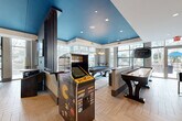 Located at 60 11th St NE in Atlanta's vibrant Midtown neighborhood, this generously sized amenity lounge spans nearly 3,000 square feet of thoughtfully designed communal space across a single floor. The open-plan layout is anchored by striking herringbone-pattern hardwood flooring that flows seamlessly throughout, tying together each distinct zone.

The entertainment-focused game room is highlighted by a bold teal-painted ceiling with recessed lighting, a full-size billiards table, a classic Pac-Man arcade cabinet, shuffleboard table, and an air hockey table—all bathed in natural light from expansive floor-to-ceiling windows overlooking the Atlanta skyline. An elegant chandelier adds a refined touch to this lively area.

A full-service bar and kitchen island anchors the social hub of the space, complete with a stainless steel prep counter, dark cabinetry, bar-height seating, and large mounted televisions. The adjacent dining and lounge area features sleek high-top tables and modern chairs beneath a curved architectural ceiling detail with slatted accents.

A dedicated media lounge showcases a textured stone accent wall displaying colorful contemporary artwork, complemented by a large flat-screen TV. Throughout, floor-to-ceiling glazing invites abundant natural light and sweeping urban views, creating a bright, energetic atmosphere ideal for both socializing and relaxation.