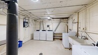 This utility room serves as a dedicated shared laundry facility, offering a practical and well-equipped space for laundry needs. The room features multiple top-loading washing machines and a row of three front-loading dryers, each vented individually through overhead ductwork that connects to the ceiling exhaust system. A double-basin utility sink is centrally positioned between the washers, providing added functionality for hand-washing or pre-treating garments. The walls are constructed of painted concrete block, giving the space a durable, no-frills character consistent with a utility environment. The floor is finished with light-colored vinyl tile, and the exposed ceiling reveals an network of pipes, conduits, and ductwork. A folding table and waste bins are available along one wall, offering a convenient surface for sorting and folding laundry. An electronic payment or control panel is mounted on the wall alongside visible electrical conduits and utility connections. A fire extinguisher is prominently installed for safety compliance. The room is accessed through a standard wood-framed door, and overhead lighting provides adequate illumination throughout the space. Overall, the room is straightforward and utilitarian, designed to efficiently serve the laundry requirements of a multi-unit building.