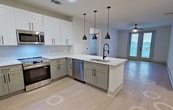 This 1,120 sq ft apartment in Charlotte, NC features 2 bedrooms, 2 bathrooms, and a well-appointed open-concept kitchen and living area. The kitchen is a standout, boasting two-tone sage green and white shaker cabinetry, white quartz countertops, a subway tile backsplash, stainless steel appliances including a French door refrigerator, range, microwave, and dishwasher, and a peninsula island with an undermount sink and matte black pendant lighting. Light wood-style flooring runs throughout the unit, complementing the clean, neutral palette. The primary bedroom spans 180 sq ft and features French doors opening to a private balcony, a ceiling fan, and access to a walk-in closet with built-in shelving and a natural light window. The primary bathroom offers a frameless glass walk-in shower with white tile surround, dual-sink vanity with sage cabinetry, white countertops, and matte black fixtures. The secondary bedroom is bright and open, served by a full bathroom with a tub/shower combo, matching vanity, and matte black hardware. Three hallways and two closets provide ample circulation and storage throughout this modern, move-in-ready Charlotte apartment.