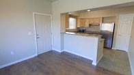 This single-story space features an open and functional layout that seamlessly connects the entryway to the kitchen area. The entryway is accented by a solid white paneled door with a clean, modern finish, opening into a living area with rich, dark hardwood-style flooring that extends throughout the main space.

The kitchen is defined by a peninsula-style breakfast bar with granite countertops featuring warm, earthy tones, providing both workspace and a casual dining option. Light wood cabinetry offers ample storage while maintaining a bright, airy feel within the kitchen. Stainless steel appliances, including a refrigerator and microwave, add a contemporary touch. A window above the sink allows natural light to filter into the kitchen, enhancing its open atmosphere.

Recessed lighting is installed in the kitchen area, providing focused illumination. The neutral beige wall tones throughout the space create a versatile backdrop that complements the warm flooring and cabinetry. White baseboards provide crisp contrast against the flooring, contributing to the overall clean and well-maintained aesthetic of the space.