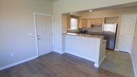 This single-story space features an open and functional layout that seamlessly connects the entryway to the kitchen area. The entryway is accented by a solid white paneled door with a clean, modern finish, opening into a living area with rich, dark hardwood-style flooring that extends throughout the main space.

The kitchen is defined by a peninsula-style breakfast bar with granite countertops featuring warm, earthy tones, providing both workspace and a casual dining option. Light wood cabinetry offers ample storage while maintaining a bright, airy feel within the kitchen. Stainless steel appliances, including a refrigerator and microwave, add a contemporary touch. A window above the sink allows natural light to filter into the kitchen, enhancing its open atmosphere.

Recessed lighting is installed in the kitchen area, providing focused illumination. The neutral beige wall tones throughout the space create a versatile backdrop that complements the warm flooring and cabinetry. White baseboards provide crisp contrast against the flooring, contributing to the overall clean and well-maintained aesthetic of the space.
