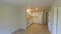 This single-floor space offers a practical and efficient layout designed for comfortable everyday living. The open-concept design connects the main living area to the kitchen, creating a sense of openness and ease of movement throughout the unit. Light wood-style laminate flooring runs continuously across the space, lending warmth and a cohesive aesthetic to the interior. The kitchen is thoughtfully arranged in an L-shaped configuration, maximizing available space while maintaining a clear workflow. White cabinetry lines the upper and lower walls, providing generous storage, while a built-in countertop with an integrated sink sits neatly along the lower cabinets. White appliances — including a refrigerator, electric range, and over-the-range microwave — complement the clean, uniform look of the kitchen. Recessed lighting in the kitchen area ensures the space is well-lit and functional. The neutral color palette of white walls and light flooring throughout the unit contributes to a bright and airy ambiance. A door visible to the right of the space suggests a separate room or entryway, adding to the overall functionality of the layout. Overall, this space presents a tidy, well-maintained environment suited for a variety of occupants.