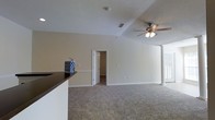 This single-floor residence features an open-concept layout that connects the living area and kitchen in a seamless, functional design. The main living space is finished with wall-to-wall carpet in a neutral tone, contributing to a warm and comfortable atmosphere. A tray ceiling adds architectural dimension overhead, while a ceiling fan with integrated light fixture serves both practical and aesthetic purposes, providing airflow and illumination throughout the room. The textured ceiling treatment complements the clean, neutral wall tones that run throughout the space.

A partial countertop — likely a kitchen peninsula or breakfast bar — separates the living area from the kitchen zone, offering a natural transition between spaces without compromising the open feel. A decorative accent piece is visible on the counter, adding a touch of personality to the otherwise minimalist interior. A white decorative column near the far corner adds a subtle architectural detail. A doorway visible from the main living area leads to an adjoining room, suggesting additional functional spaces beyond. Ample wall outlet placement throughout indicates versatile furniture arrangement possibilities. Natural light filters in from a window, keeping the space bright and inviting.