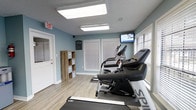 This single-floor fitness room offers a practical and inviting space designed for residents or guests seeking convenient exercise options. The room is finished with soft blue walls and light wood-look flooring, creating a clean and energizing atmosphere. Bright flush-mount ceiling panels provide ample illumination throughout the space, complementing the natural light that filters in through multiple windows dressed with white horizontal blinds. The room is equipped with several cardio machines, including treadmills and an elliptical, arranged to make efficient use of the available floor area. A wall-mounted flat-screen television is positioned for easy viewing during workouts, adding to the overall functionality of the space. An open wood shelving unit provides additional storage for accessories or personal items. The room features a clearly marked exit door, ensuring safety and ease of access. The overall design is straightforward and purposeful, prioritizing usability and comfort in a well-maintained environment suited for a focused fitness routine.