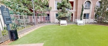 This meticulously designed outdoor space is a central feature of a multi-story residential complex in Irvine, California. The courtyard boasts a large, well-maintained artificial turf area, providing a lush green centerpiece that remains pristine year-round. The space is enclosed by decorative metal fencing, which offers both security and aesthetic appeal.

Strategically placed white benches provide comfortable seating areas for residents to relax and enjoy the outdoors. Tall pine trees and palm trees surround the courtyard, creating a natural backdrop and offering partial shade. The landscaping includes a variety of plants and shrubs, adding texture and color to the environment.

The building's architecture features a soft pink exterior with large, arched windows, creating an elegant backdrop for the courtyard. Outdoor lighting fixtures are installed throughout the space, ensuring the area is well-lit and inviting during evening hours.

This thoughtfully planned outdoor area serves as a communal gathering space, perfect for relaxation, social interactions, or quiet contemplation. It seamlessly blends natural elements with modern residential living, providing residents with a serene escape within their urban environment.