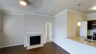 This single-floor residence offers a thoughtfully designed interior that balances classic detailing with a warm, inviting atmosphere. The living area serves as the heart of the home, anchored by a fireplace with a white wood mantle and elegant marble tile surround — a standout architectural feature that adds both charm and character to the space. Rich, dark hardwood floors extend throughout, providing a striking contrast against the neutral, light-toned walls and crisp white trim work. Crown molding runs along the ceiling line, lending the space a refined and polished finish. A ceiling fan adds functional comfort overhead. The open layout connects the living area to the kitchen, where a granite countertop overhang is visible, hinting at a bar-style seating area ideal for casual dining or entertaining. The kitchen's adjacency to the main living space enhances the overall sense of openness and flow. A paneled interior door leads to an additional room, offering connectivity between the living spaces. Natural light filters into the space, brightening the neutral palette and highlighting the quality of the finishes throughout. The overall ambiance is clean, classic, and move-in ready, with traditional architectural details that give the space a timeless appeal.