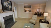 This single-floor residence showcases an open-concept design that seamlessly connects the living, kitchen, and dining areas. The living room serves as the focal point, anchored by a classic white-mantled fireplace with a dark tile surround, adding both warmth and architectural character to the space. Neutral-toned carpeting flows throughout, complementing the warm beige walls adorned with crisp crown molding that lends a refined finishing touch.

The kitchen is accessible via a wraparound peninsula counter, creating a natural pass-through that maintains visual connectivity with the rest of the living space. It features light wood cabinetry, a stainless steel refrigerator, and a contrasting dark upper accent cabinet. An under-cabinet fluorescent light provides task illumination over the work surfaces.

Adjacent to the kitchen, the dining area is defined by a elegant chandelier that draws the eye and distinguishes the space. A set of dark wood dining chairs surrounds the table, creating a welcoming setting for meals. Together, the rooms form a cohesive, functional layout bathed in a neutral palette that lends the home a calm, inviting ambiance.