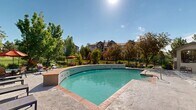 Located at 1495 Farnham Pt in Colorado Springs, Colorado, this single-level outdoor amenity space centers around a generously sized freeform swimming pool with striking turquoise water and brick tile edging. The pool area is complemented by an adjacent rectangular pool or spa section near the main clubhouse building, which features a dramatic arched window facade that allows abundant natural light into the interior. A broad, stamped concrete sun deck surrounds the pool, furnished with cushioned lounge chairs positioned to take full advantage of Colorado's abundant sunshine. A covered outdoor pavilion or pergola structure sits adjacent to the main building, offering a shaded retreat for residents. The grounds are beautifully landscaped with mature deciduous trees, ornamental shrubs, and native plantings, providing both privacy and natural beauty throughout the seasons. A black iron perimeter fence encloses the entire pool area, ensuring safety while maintaining open sightlines to the surrounding greenery and residential buildings beyond. Red umbrella accents and additional lounge seating are distributed around the deck, creating a welcoming, resort-style atmosphere. The space benefits from expansive views of the landscaped community grounds and Colorado's clear blue skies, making it an ideal outdoor gathering and relaxation destination.