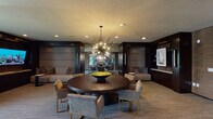 This refined, single-floor space presents a seamlessly curated blend of contemporary design and upscale functionality. The main lounge area is anchored by a substantial round dark wood table surrounded by modern upholstered chairs with brass-toned frames, all positioned beneath a striking Sputnik-style chandelier that serves as a dramatic focal point. Recessed lighting throughout the smooth ceiling complements the ambient warmth of the space.

The interior is defined by rich, dark wood wall paneling paired with textured woven wall treatments, creating a layered and sophisticated palette. Built-in shelving and display areas add a polished, purposeful character to the walls. Full-length glass partitions with horizontal louvered blinds divide the space into distinct zones, allowing visual connectivity while providing privacy. These partitions separate the central lounge from additional seating areas and what appears to be a fitness or wellness room visible through the glazed walls.

A cozy lounge alcove features a large, upholstered built-in sofa framed by a dark architectural surround with integrated lighting, offering an intimate setting alongside a side table and large-format display screen. Adjacent seating areas with armchairs and natural light from full-height windows with draped curtains further enhance the openness. Entry points feature elegant dark-framed glass doors set within textured wall surrounds, completing the space's refined and professional atmosphere.